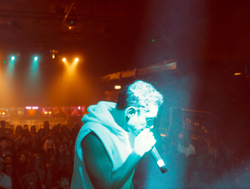 Mogul Mowgli: Riz Ahmed raps in front of a crowd, bathed in a blue spotlight and wearing a sleeveless white hoodie.